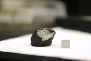 The largest of the fifteen meteorites next to a one-centimeter cube for scale. The black outer layer is called a “fusion crust” and forms when the outside melts as the objects hurtles through the atmosphere at high velocity. (Photo by Anna Copper/Cronkite News)